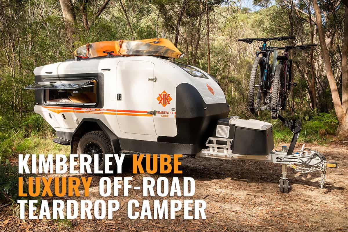 A Kimberley Kube teardrop camper trailer parked on grass with two bicycles on a rear rack and a kayak mounted on top, set against a backdrop of trees and a clear sky.