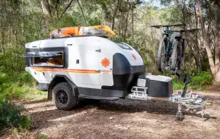 Teardrop camper with iSi bicycle rack attached, parked outdoors with Australian bush background. Passenger side view.